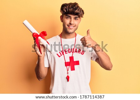 Young hispanic man wearing lifeguard t shirt using binoculars smiling happy and positive, thumb up doing excellent and approval sign 