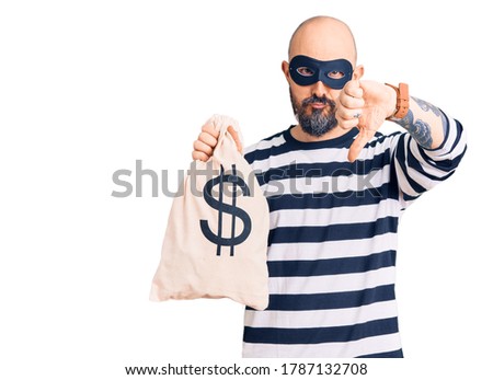 Young handsome man wearing burglar mask holding money bag with dollar symbol with angry face, negative sign showing dislike with thumbs down, rejection concept 