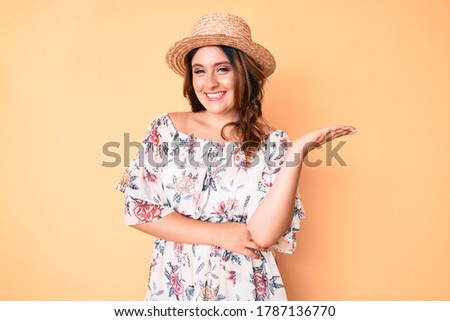 Young beautiful caucasian woman wearing summer dress and hat smiling cheerful presenting and pointing with palm of hand looking at the camera. 