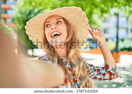 Young beautiful blonde woman on vacation wearing summer hat smiling happy. Standing with smile on face making selfie by the camera at street of city.