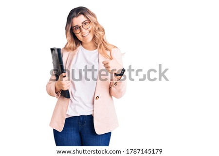 Young caucasian woman wearing business clothes and glasses holding binder smiling happy and positive, thumb up doing excellent and approval sign 
