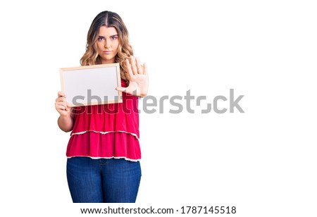 Young caucasian woman holding empty white chalkboard with open hand doing stop sign with serious and confident expression, defense gesture 