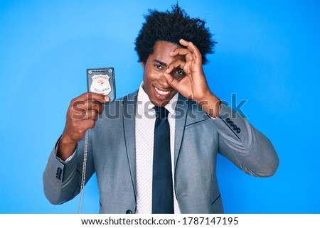 Handsome african american man with afro hair holding detective badge smiling happy doing ok sign with hand on eye looking through fingers 