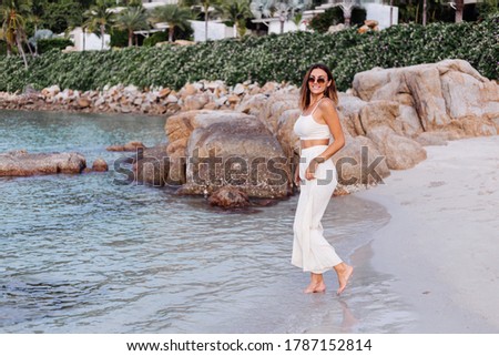 Romantic portrait of young calm happy caucasian fit slim woman in crop cami top and pants set alone on rocky tropical beach at sunset. 