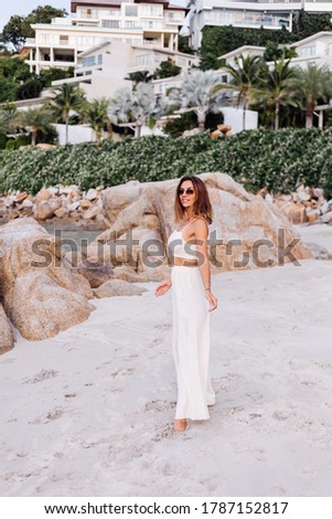 Romantic portrait of young calm happy caucasian fit slim woman in crop cami top and pants set alone on rocky tropical beach at sunset. 