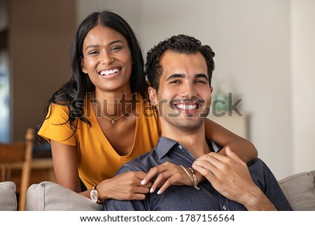 Smiling indian woman hugging her husband on the couch from behind at home. Loving middle eastern couple looking at camera with big grin. Portrait of laughing girl embracing handsome latin man on sofa.