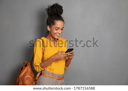 Young beautiful african woman with leather backpack standing on grey wall while using smartphone. Stylish latin girl isolated against gray background checking mobile phone with copy space.