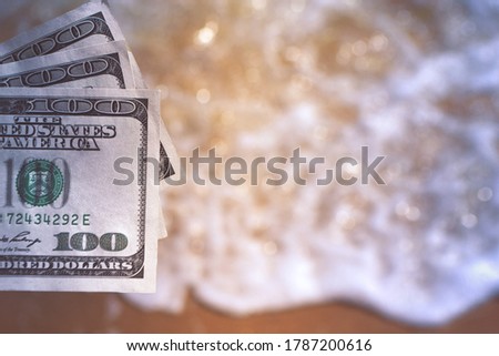 Girl holding money bill of 300 dollars on background of sea ocean waves with white foam and sand wet beach close-up. Hand wave sea ocean money dollars vacation. Concept finance money holiday traveling