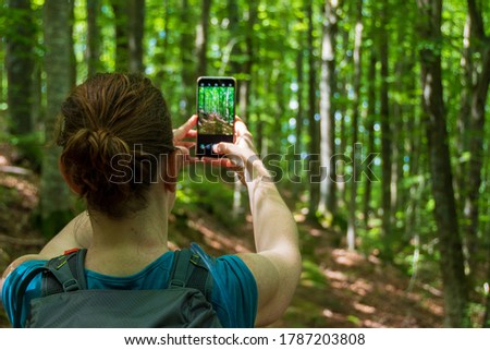Young woman with a backpack hiking alone on a trail in a shaded green forest stops to take a picture with her phone. Orecchiella Natural Reserve - San Romano in Garfagnana, Tuscany - Italy