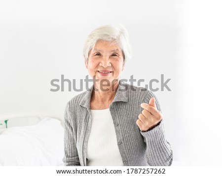 old asian patient sitting on bed, she feeling happy and smile, she show mini heart sign with hand in hospital, elderly healthcare