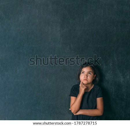 Back to school. Serious little girl thinking touching her head with a finger near blackboard. Child from elementary school. Education.