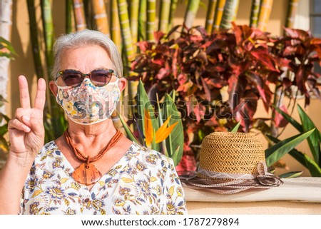 Senior woman wearing floral mask due to coronavirus making ok sign with hands standing against a tropical garden - concept of active elderly and positive vibes
