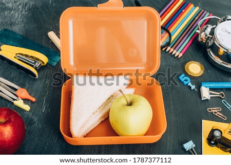 top view of lunch box with sandwiches and ripe apple near school stationery on black chalkboard