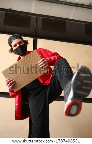 A young man of 25-30 years old in a black protective mask, yellow glasses, a cap and a red jacket holds an empty cardboard sign in his hands. Protest concept. Blank mock up for design, mockup