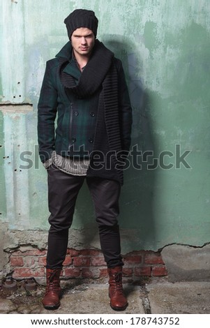 full length picture of a young fashion man posing in front of a cracked wall with his hands in his pockets and an angry look on his face