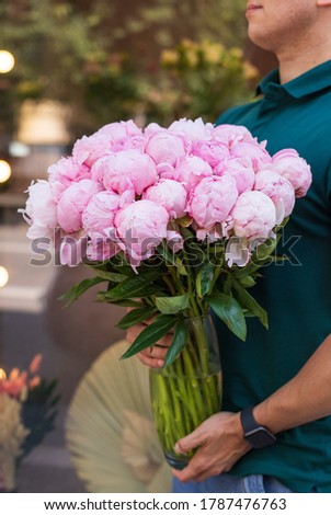 Young man florist holding big beautiful blossoming mono bouquet of pink peonies in glasses vase.