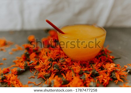 A small cup is filled to the top with yellow liquid honey. Medicinal herbal dried plants marigold, orange calendula. Neutral white and gray background.
