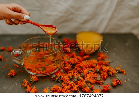 A woman's hand pours honey from a spoon into a cupFlower tea in a large transparent mug.A small cup is filled to the top with yellow liquid honey.Medicinal herbal dried plants marigold.Gray background