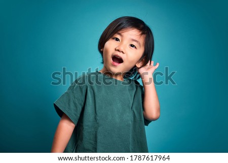 Exited asian little kid is listenning a secret or paying attention to sound. Empty space in studio shot isolated on colorful bue background. Education concept for school.