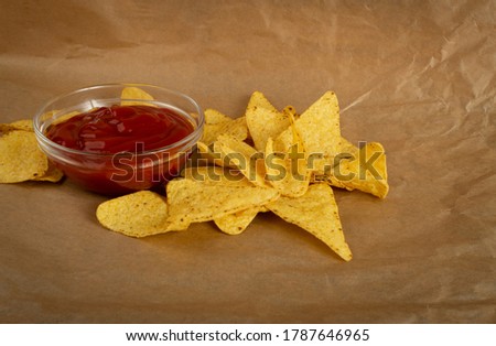 Triangle corn chips with red sauce on rustic background. Mexican nachos chips for nacho tortilla, maize snack, corn crisps or totopos with salsa dip on crumpled craft paper
