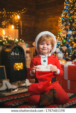 Child with a christmas socks on wooden background. Happy little smiling boy is wearing Santa clothes. Happy kid having fun with gift. Christmas tree bachground