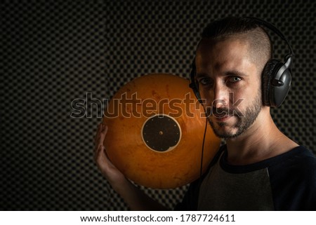 Dj with blue eyes poses with an orange record and headphones, dressed in a gray shirt with navy blue sleeves, inside a recording studio