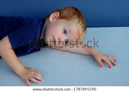 Portrait of a charming blond boy with blue eyes. A three-year-old boy lies on his side and looks into the distance. Sad child.