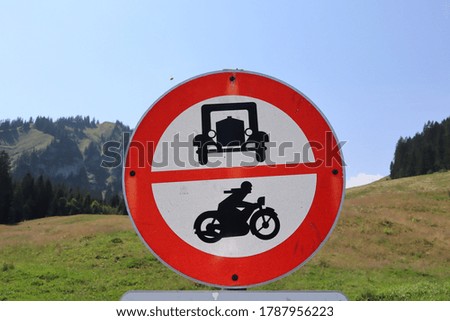 Round red and white do not enter sign with black silhouette depictions of a very old fashioned car and motorcycle. A meadow and green mountains are in teh background.
