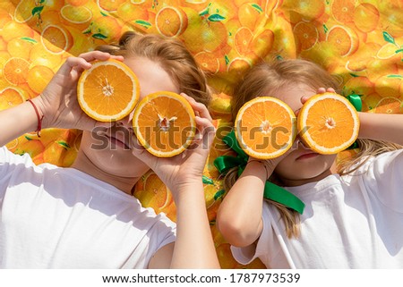 Two girls sisters with oranges in hands are lying on orange blanket on grass outdoors. Summer bright colors, healthy food, vitamins and trace elements. Photo of advertising concept of fruit juices