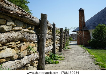A path along an old stone wall on one side and a green lawn on the other, leading to a white house with a large brick chimney and a tapered roof. A hot summer sunny day and a beautiful blue cloudless 