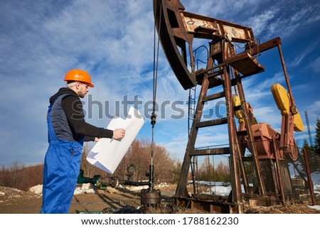 Side view of oil worker holding plan of oil field. Engineer in work uniform and helmet standing near petroleum pump jack under beautiful cloudy sky. Concept of petroleum industry and oil extraction.