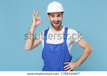 Cheerful young man in coveralls protective helmet hardhat isolated on blue background studio portrait. Instruments accessories for renovation apartment room. Repair home Showing Ok gesture Royalty-Free Stock Photo #1788166847