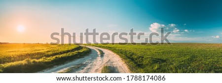 Sun Sunshine In Sunset Bright Sky. Beautiful Evening Sky Above Rural Landscape With Country Road. Young Green Wheat Field Meadow And Countryside Road. Agricultural And Weather Forecast Concept Royalty-Free Stock Photo #1788174086