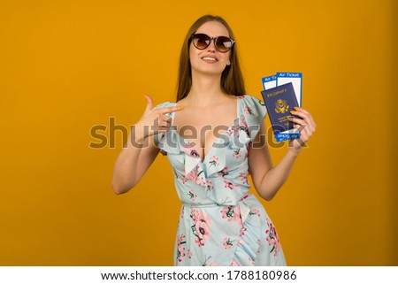 Joyful young woman in blue dress with flowers and sunglasses pointing to airline tickets with a passport on a yellow background. Rejoices in the resumption of tourism after the coronovirus pandemic.