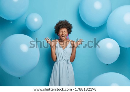 Shot of upbeat cheerful dark skinned woman feels very happy and excited, raises palms and laughs, spends free time on party, wears nice blue summer dress with earrings and rings, poses near balloons