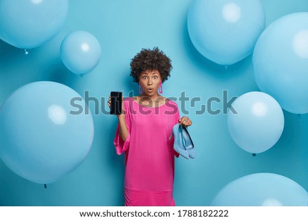 Photo of astonished curly young woman shows mobile phone display and high heeled shoes, makes shopping online, buys clothes in internet store, stands against blue background with balloons around
