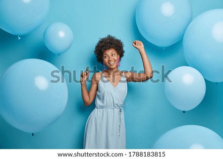 Happy dark skinned woman enjoys music at party, dances carefree, has fun and moves with rhythm of merry song, dressed in festive outfit, isolated over blue background with decorated air balloons.