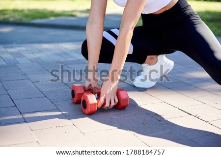 Close-up picture of female hands, holding red fitness weights, doing power morning exercises in city park in summer. Training process outdoors. Close-up image of woman's body, stretching.
