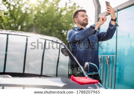 Young smiling handsome bearded man in jeans shirt, standing near his new luxury electric car and taking selfie pictures with his smartphone. Happy satisfied new owner of the car outdoors