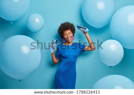Pretty curly woman chills at party, dances happily, keeps hands raised with shoes, spends time in night club, takes off high heel shoes, poses against blue wall. Monochrome shot. Holiday, celebration Royalty-Free Stock Photo #1788226415