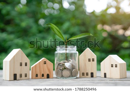 Coins are in the jar and there are plant growing in the jar. There are wooden houses all around. Saving means to buy a house for growth.Concept saving money, growth, economic business and investment.