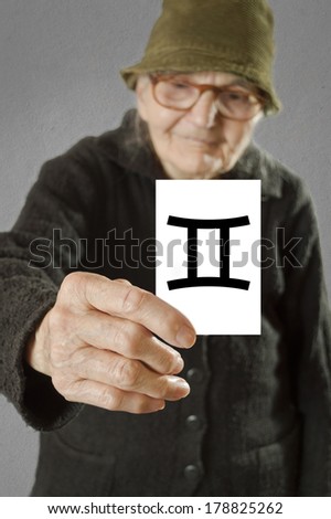 Elderly woman holding card with printed horoscope Gemini sign. Selective focus on card and fingers.