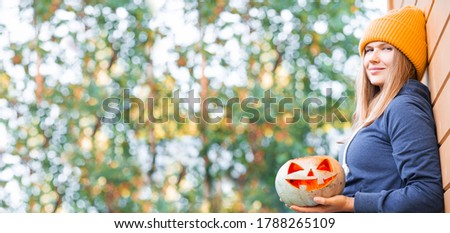 Woman in casual clothes and hat holding Halloween pumpkin over autumn nature background with copy space for text