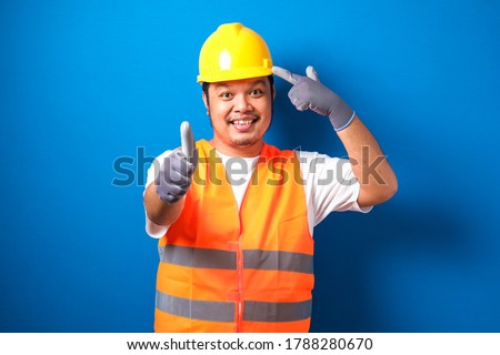 Asian construction worker man wearing orange safety vest and helmet over blue background smiling pointing to head with his finger, and other hand show thumb up as a good sign. great idea or thought
