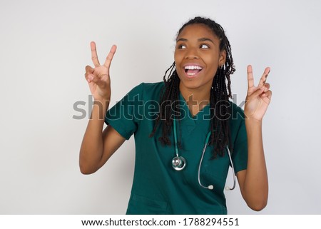 Isolated shot of cheerful doctor woman makes peace or victory sign with both hands, dressed in medical uniform, feels cool has toothy smile, isolated over gray background. People and body language.