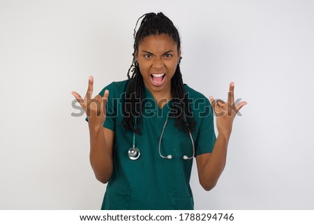 Born to rock this world. Joyful doctor woman in medical uniform screaming out loud and showing with raised arms horns or rock gesture, expressing excitement of being on concert of band.