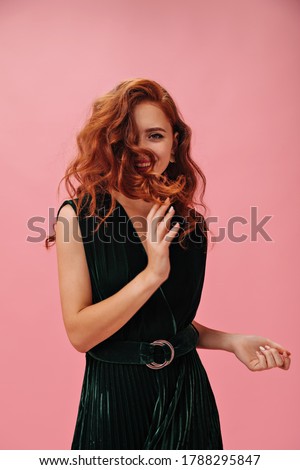Happy Woman in green outfit plays hair on pink background. Charming red haired lady in dark stylish dress has fun on isolated backdrop.. Royalty-Free Stock Photo #1788295847