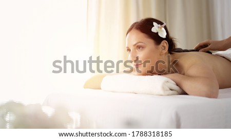 Pretty brunette woman enjoying treatment with hot stones in sunny spa salon. Beauty concept