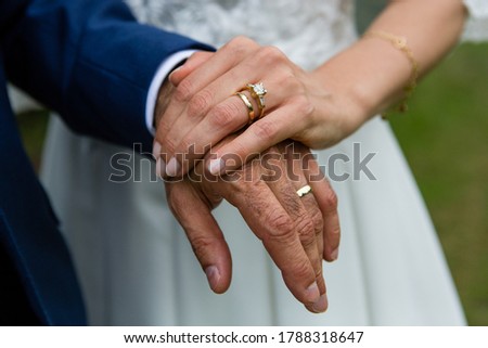 Close up picture of the bride's hand with engagement ring on the finger,  elderly groom's hand, golden wedding rings on the fingers, holding together, navy groom's suit, white bridal dress