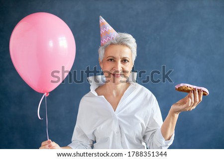 Horizontal image of stylish attractive middle aged businesswoman with pixie hairdo holding dessert and air balloon having fun at birthday party, celebrating 50th anniversary. People and aging concept